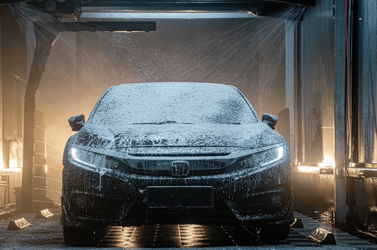 A black Honda sedan covered in white soap foam, parked inside a car wash tunnel with warm lights and water spray visible.